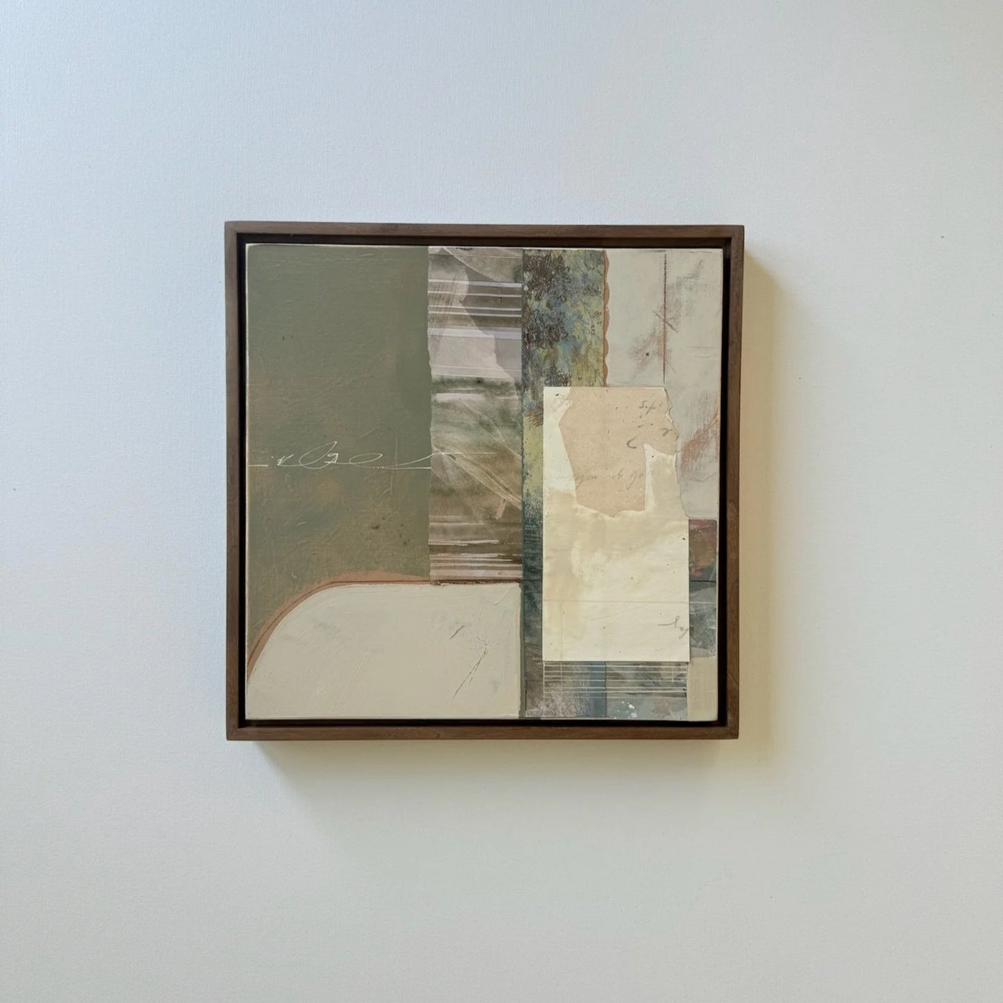 A framed abstract mixed-media collage painting hanging on a light gray wall. The artwork features a dark brown wooden frame and contains blocks of muted olive green, beige, cream, gray, and textured paper details.