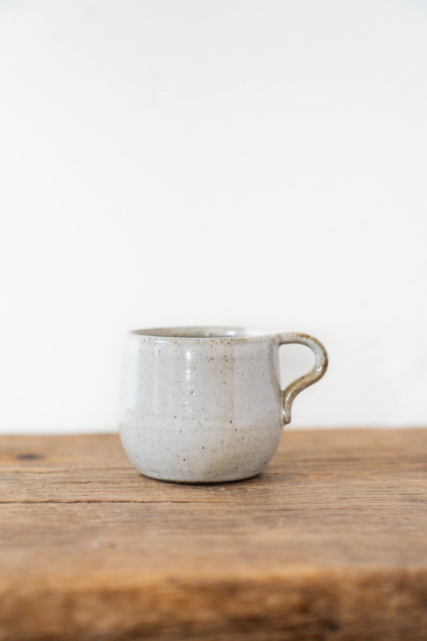 A small handmade ceramic mug with a speckled light gray-beige glaze and a rustic brown handle, sitting on a wooden surface against a plain white background.