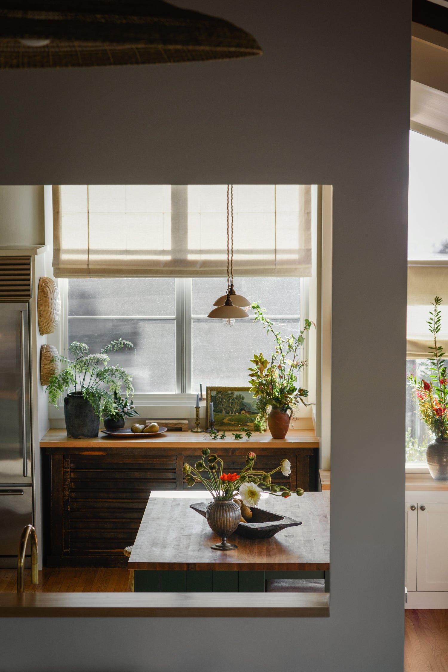 A rustic kitchen interior with a dark wood island, potted green plants, a window with beige roller shades, and a hanging pendant light. The scene is viewed through a doorway, with a small vase of flowers on the island in the foreground.