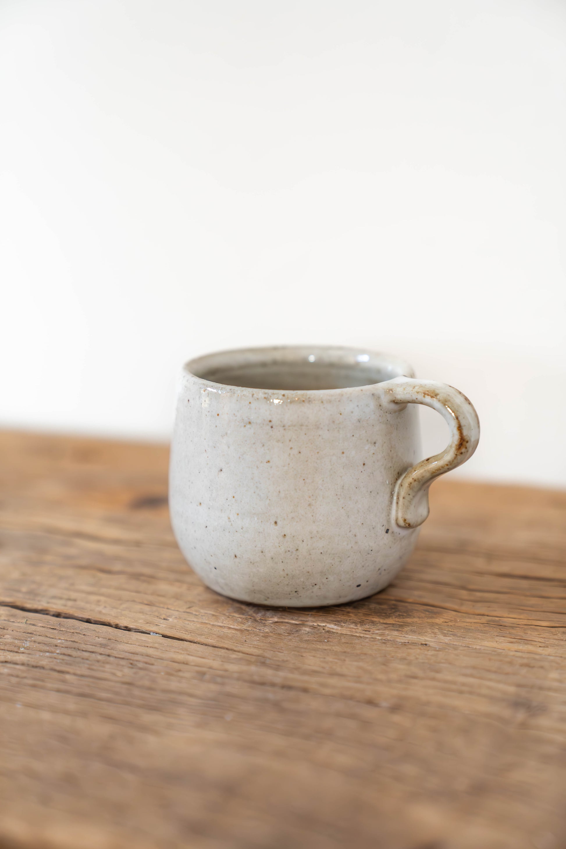 A small handmade ceramic mug with a rounded body and a unique loop handle, resting on a rustic wooden surface against a plain white background.