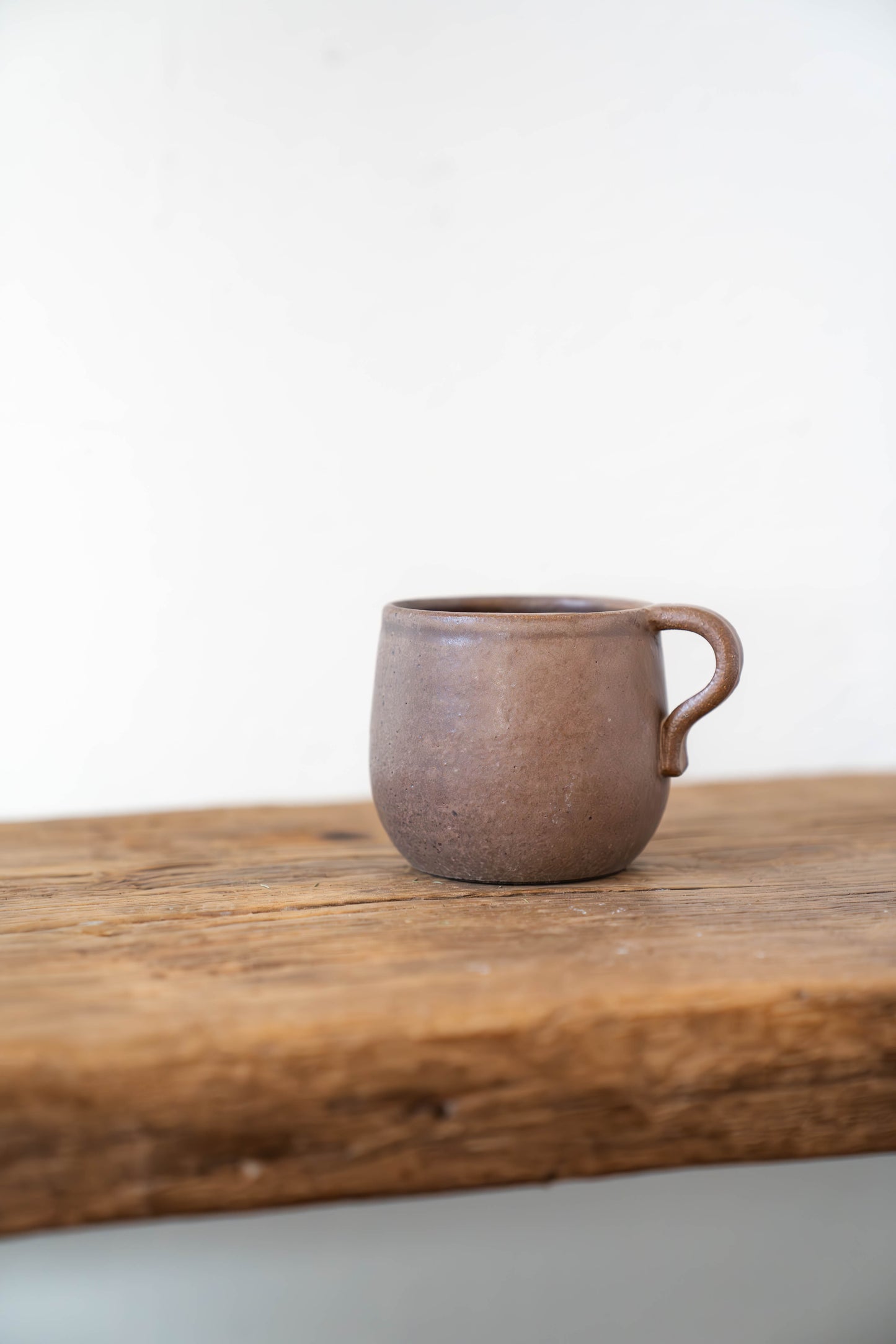 A small handmade ceramic mug with a handle, featuring a textured earthy brown glaze, resting on a rustic wooden shelf against a plain white wall.
