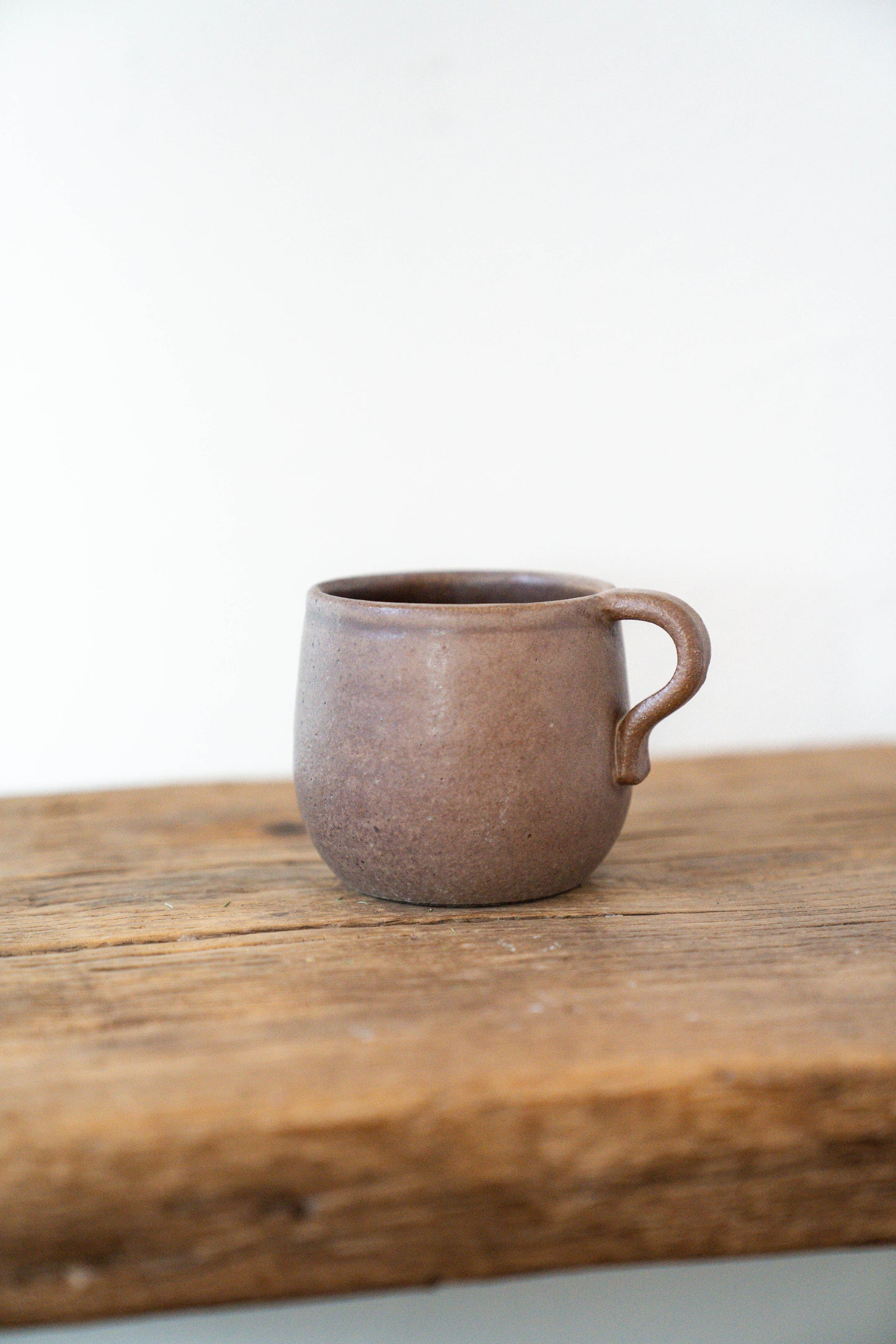A small handmade ceramic mug with a rounded body and a loop handle, finished in a textured earthy brown glaze, resting on a rustic wooden surface against a plain white background.