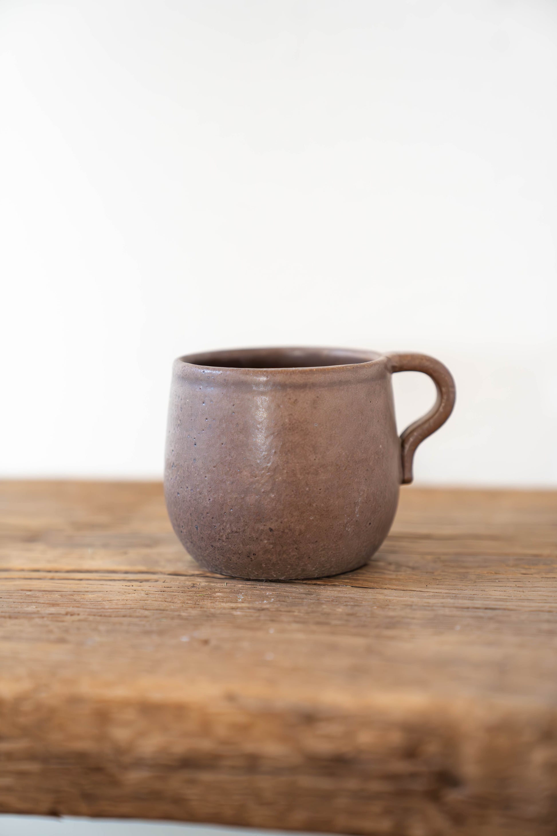 A small rustic ceramic mug with a handle, featuring a textured earthy brown glaze, sitting on a weathered wooden surface against a plain white background.