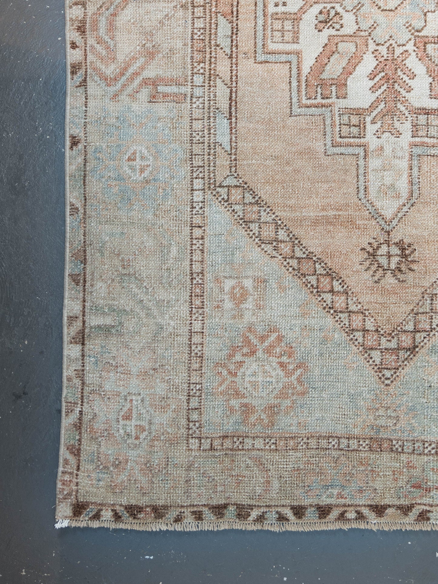 A close-up of a vintage-style area rug corner, showing a faded geometric pattern in beige, dusty blue, and terracotta brown on a dark gray floor.