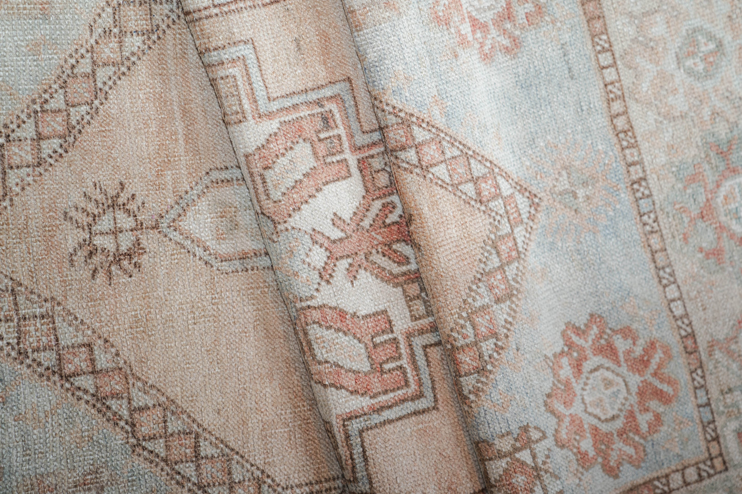 A close-up of a folded patterned textile, likely a rug or fabric, showing a distressed geometric design in muted beige, dusty pink, and light blue-gray tones.